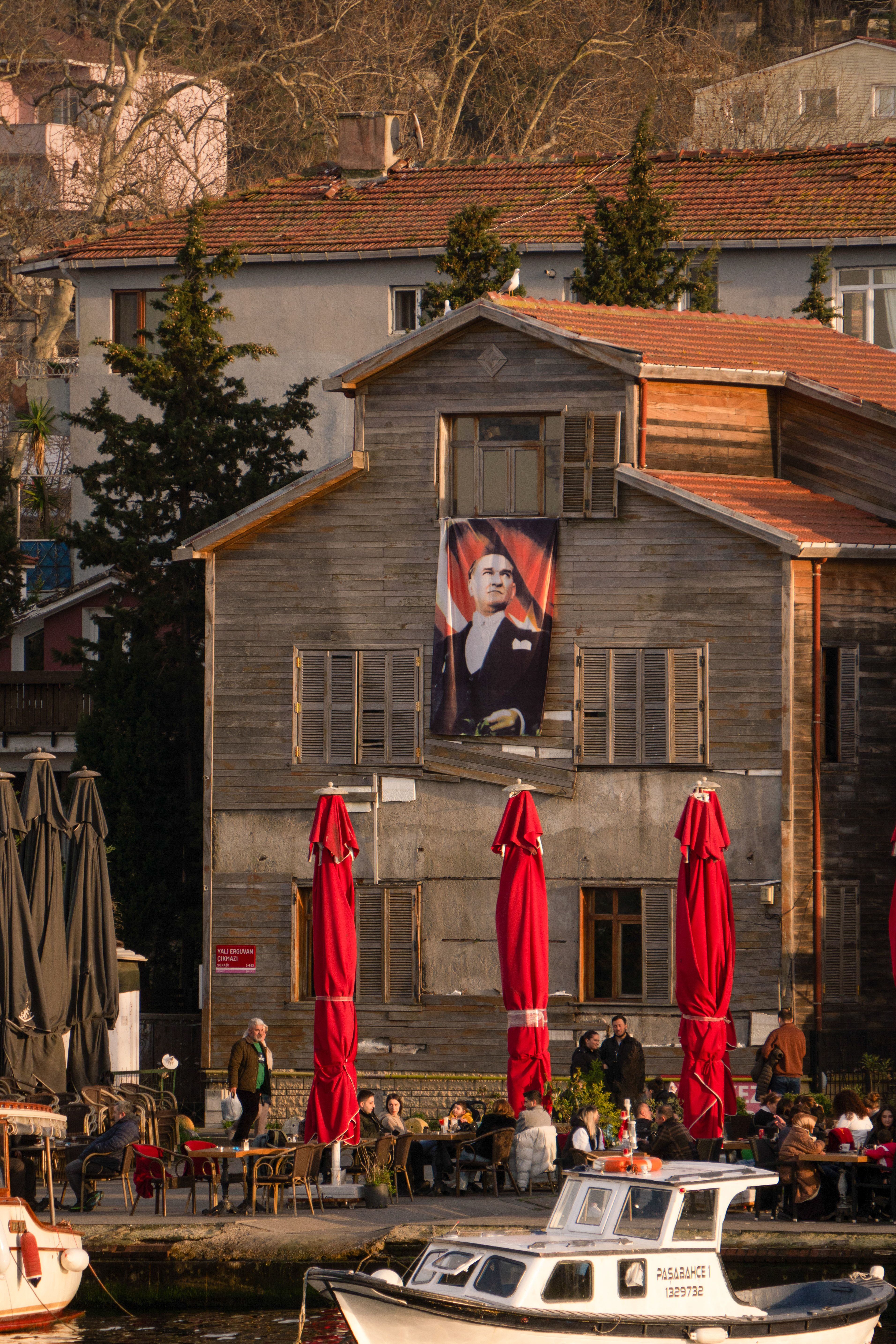 Charming café with red umbrellas by the pier in a Turkish town, showcasing vibrant urban life.