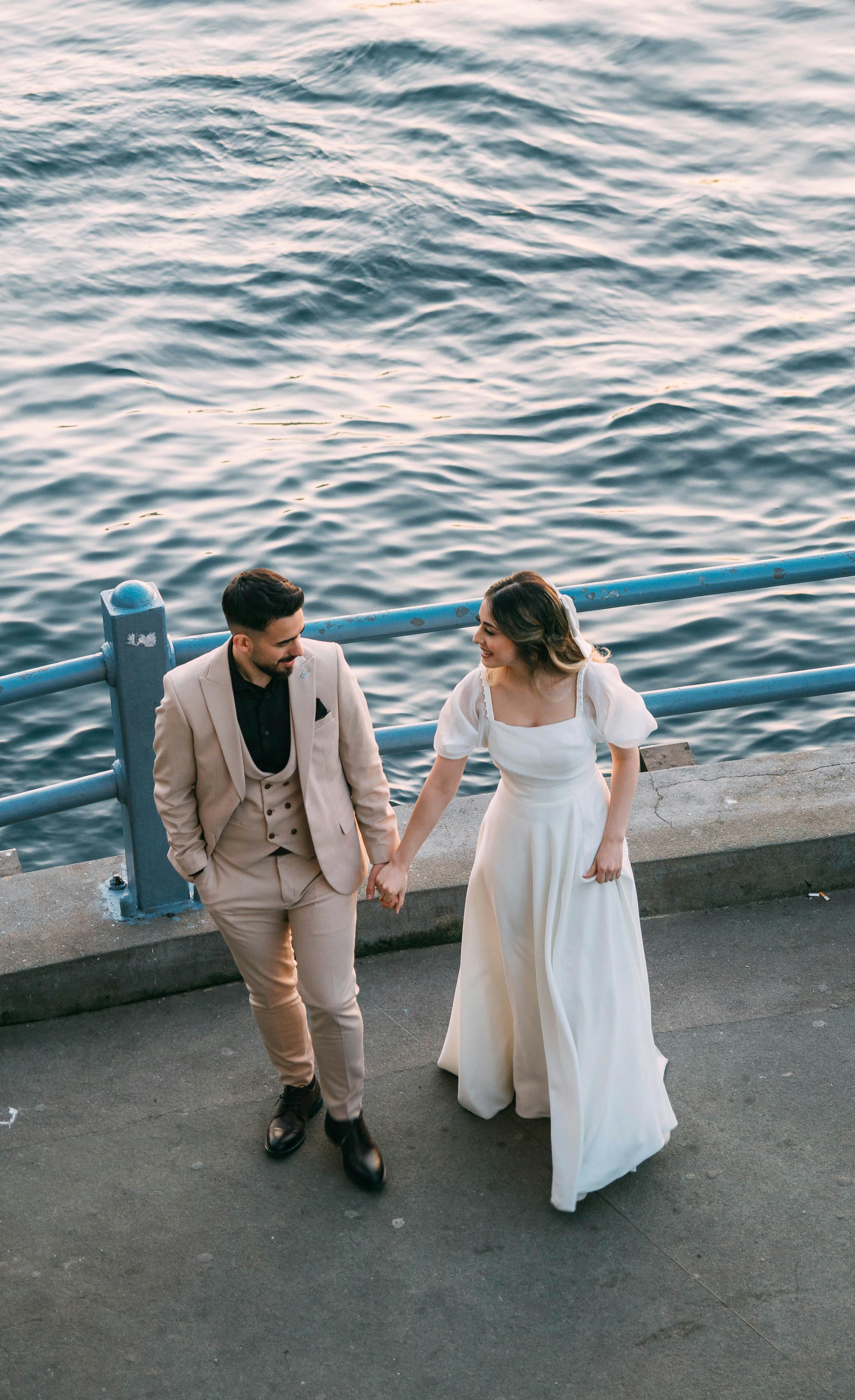 A newlywed couple walks hand in hand by the seaside in elegant wedding attire.
