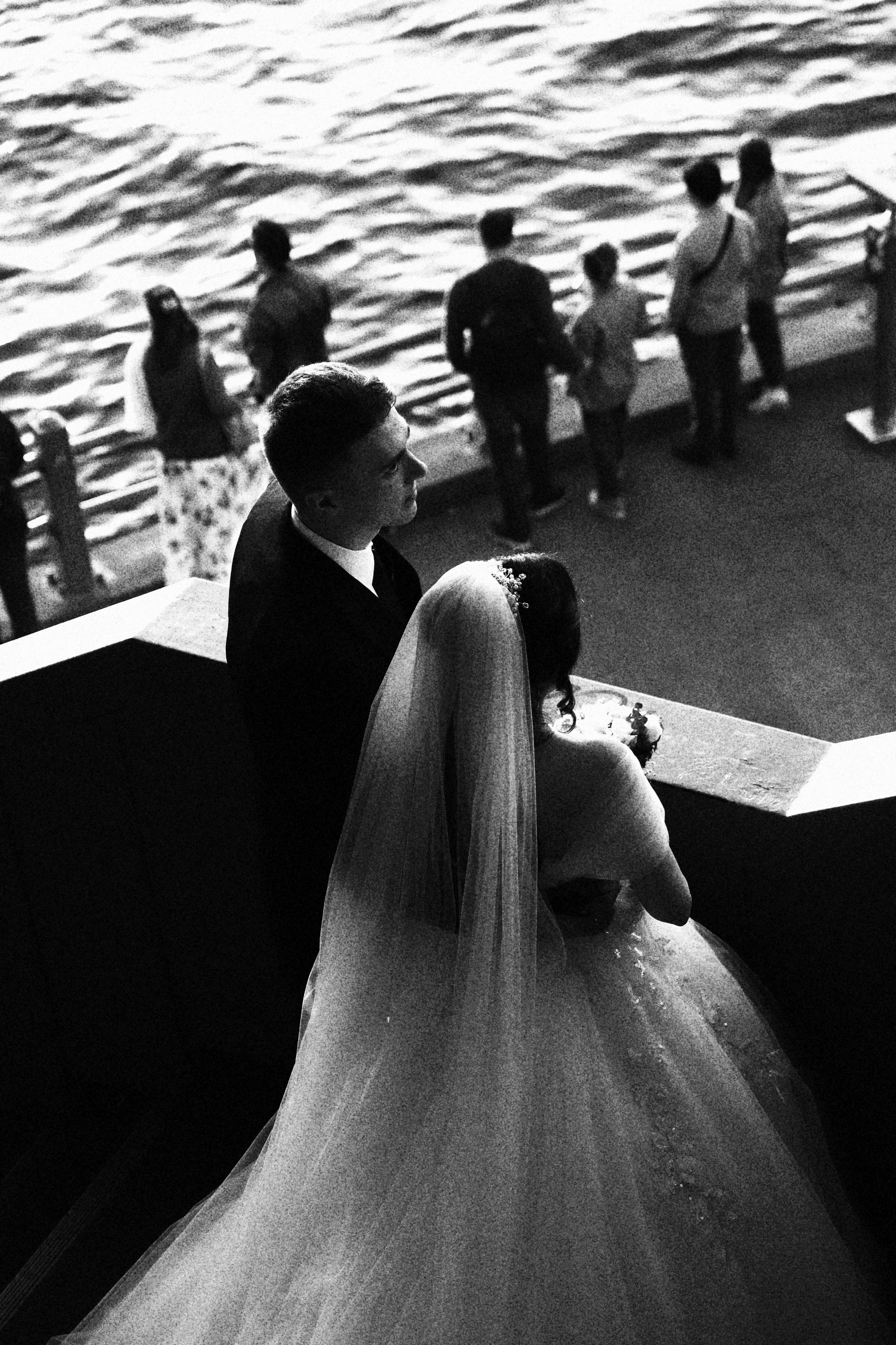 Newlyweds gazing at the sea from a city promenade, captured in black and white.