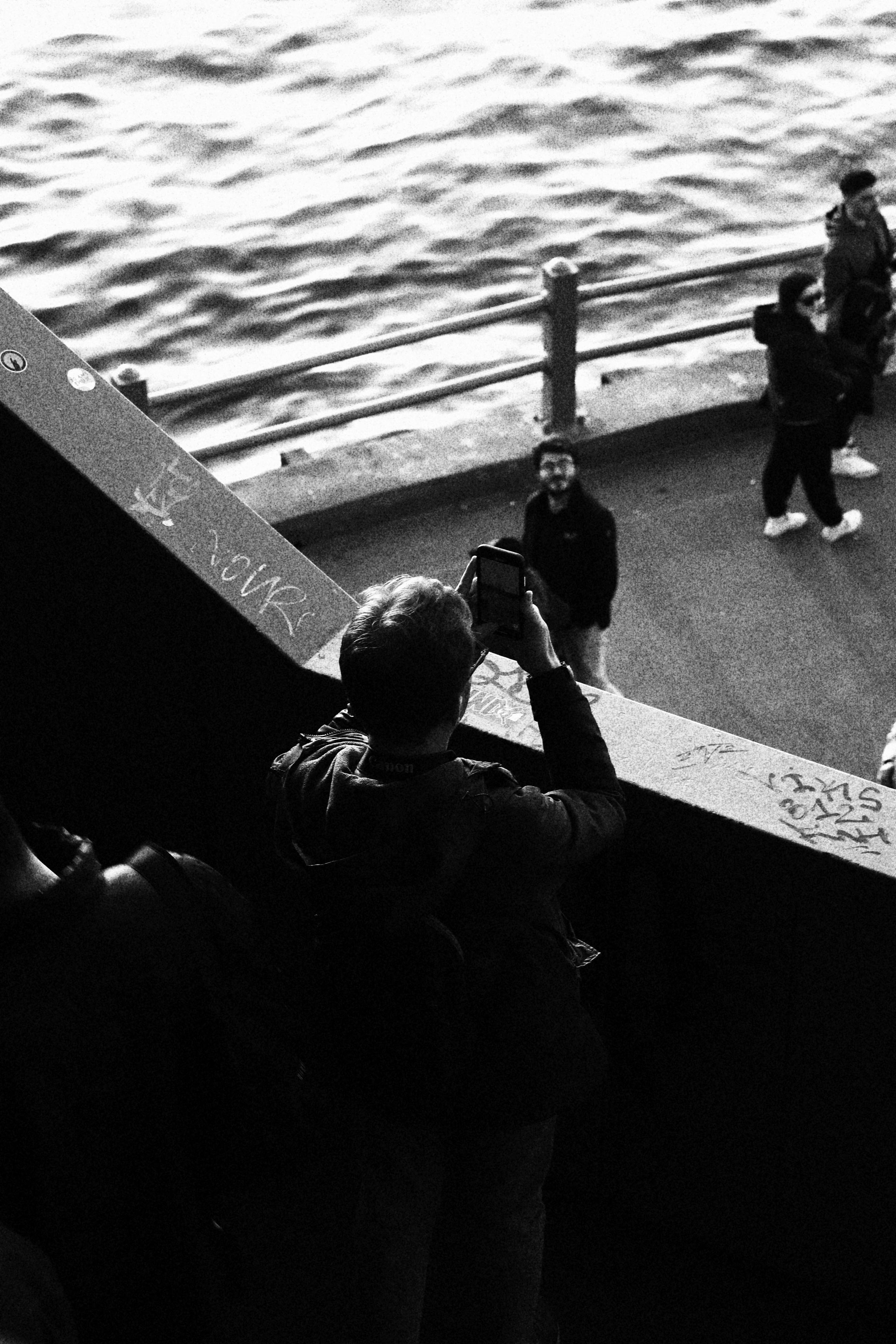 Monochrome image of tourists at a seaside promenade, capturing moments on smartphones.