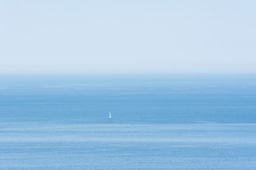 Free A lone sailboat glides over a vast, serene blue ocean under a clear sky. Stock Photo