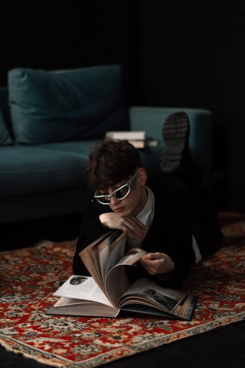 Man in Sunglasses Lying Down on Carpet and Reading Book