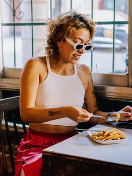 Woman in Buenos Aires diner savoring fries, wearing sunglasses, and showing tattoos.