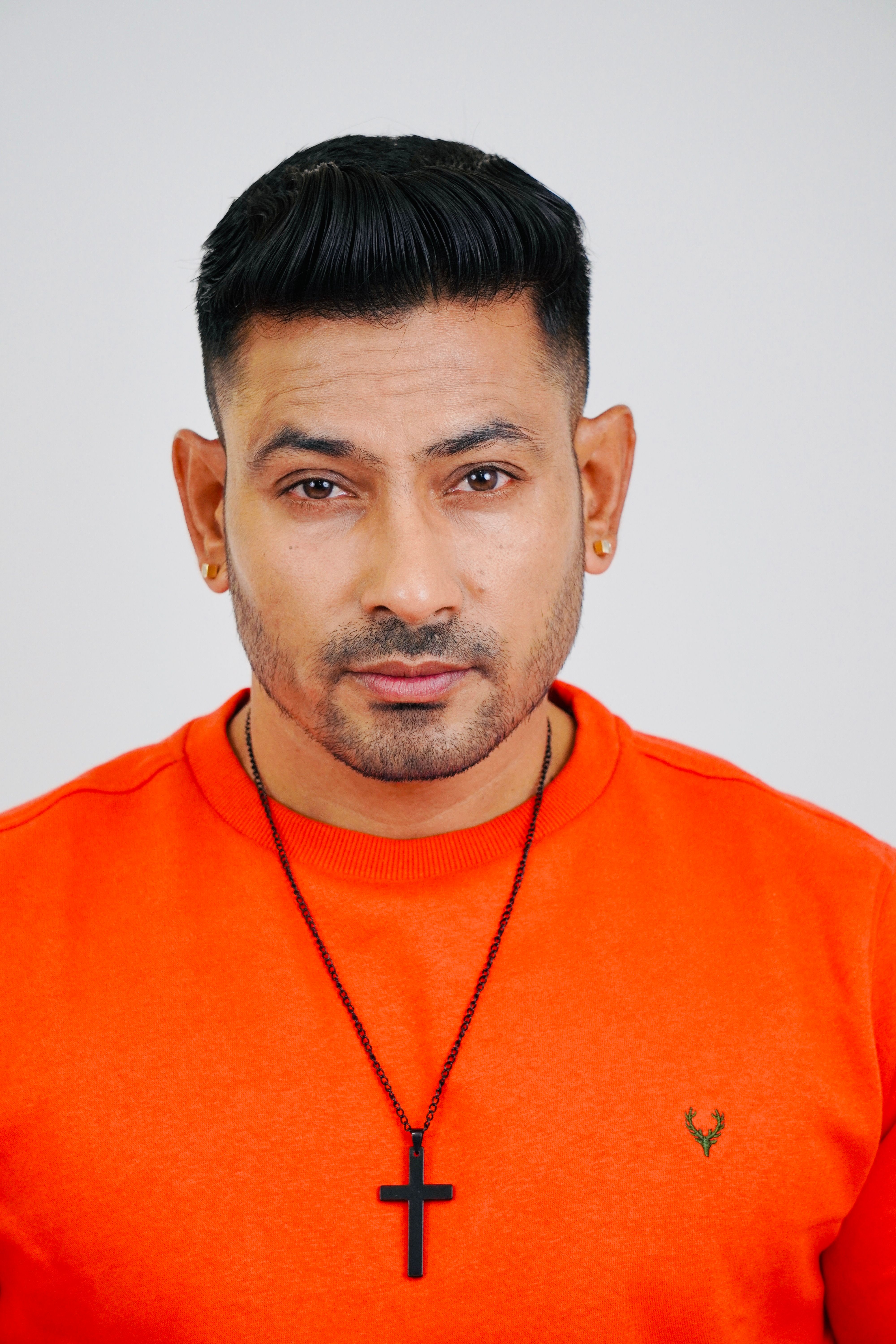 Portrait of a man wearing an orange sweater and cross necklace, looking directly at the camera.