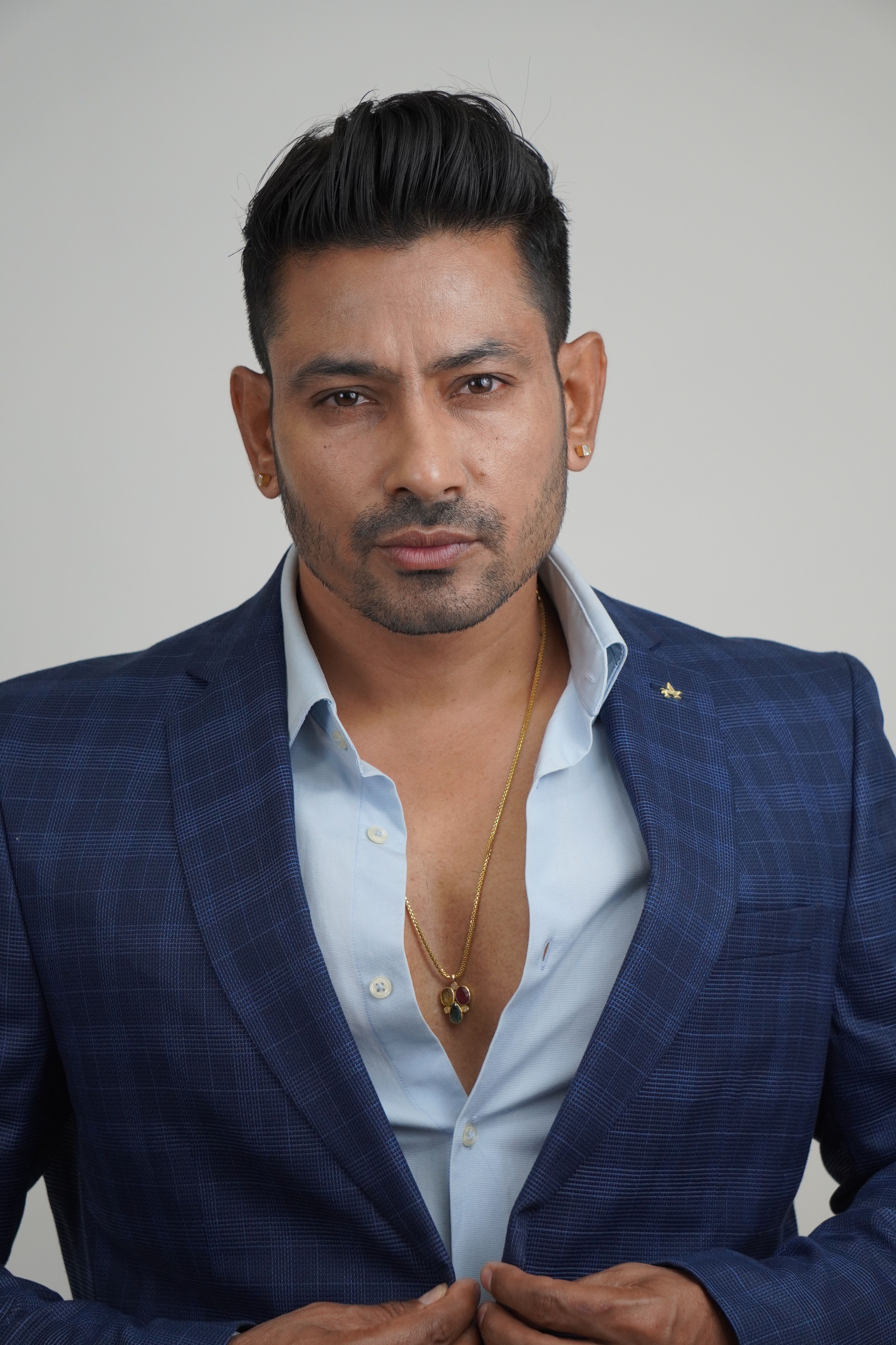 Portrait of a confident man in a blue suit with a necklace, captured in studio lighting.