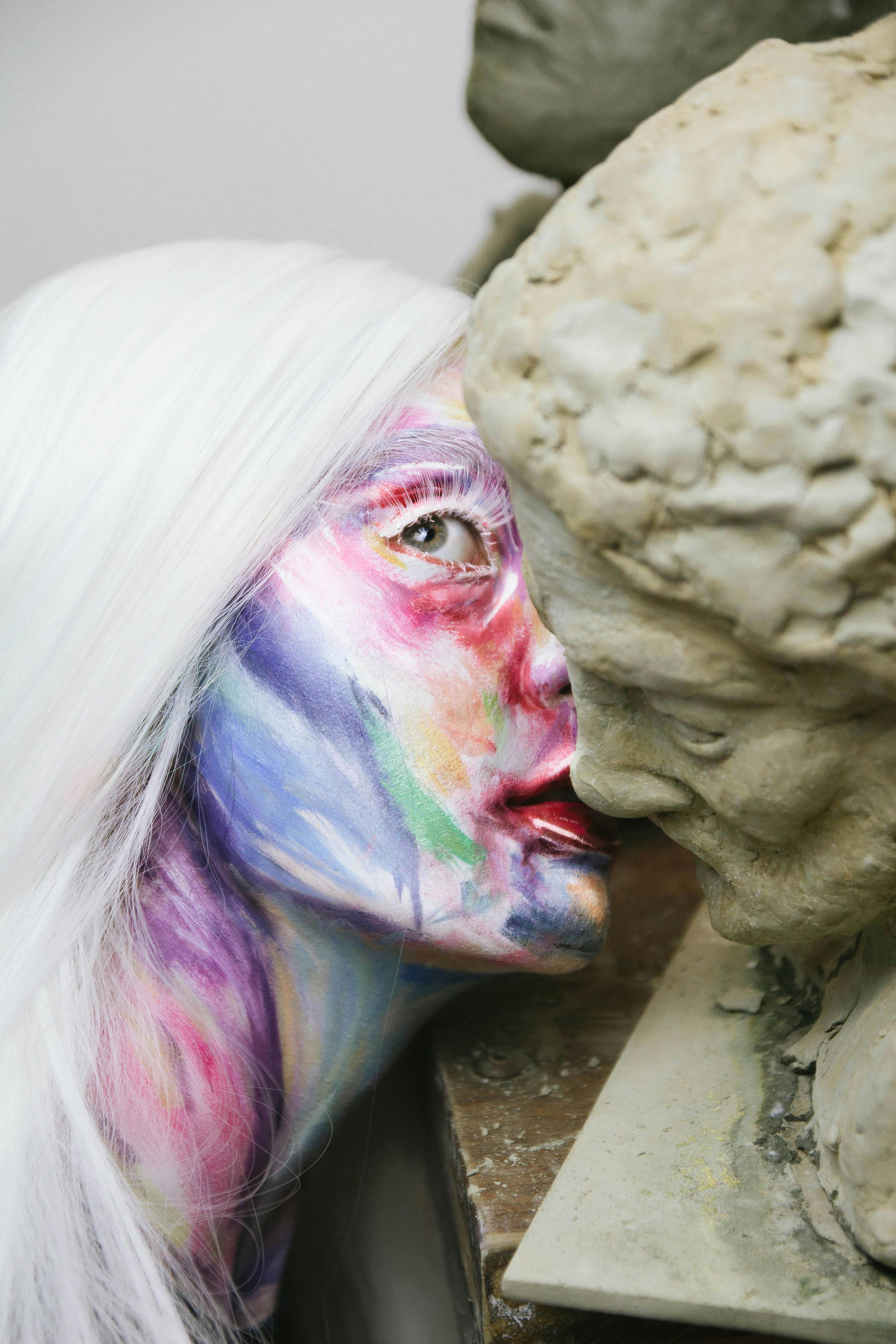 A woman with vibrant face paint interacting closely with a clay sculpture indoors.