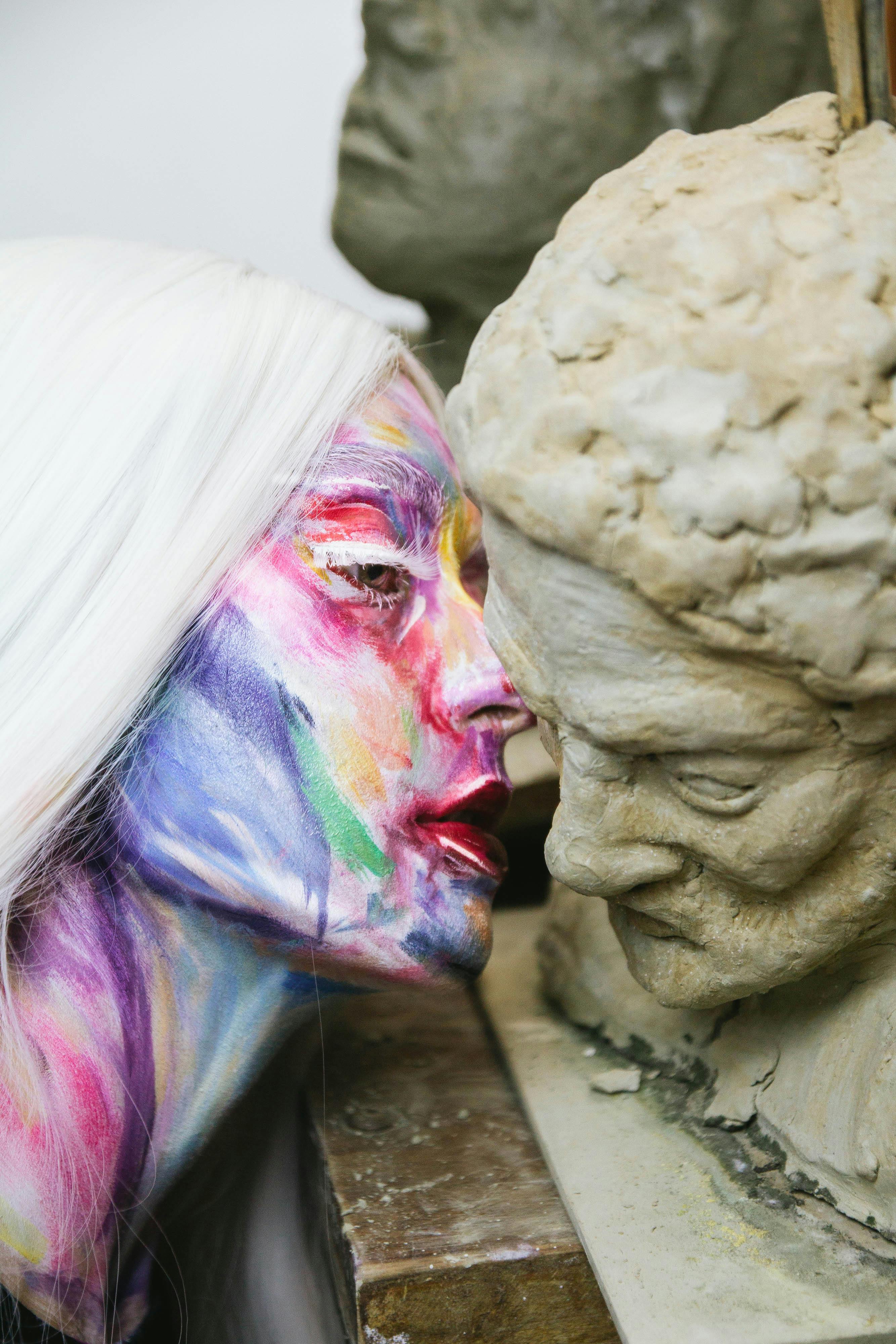 Vibrant painted face of woman interacting closely with clay sculpture bust.