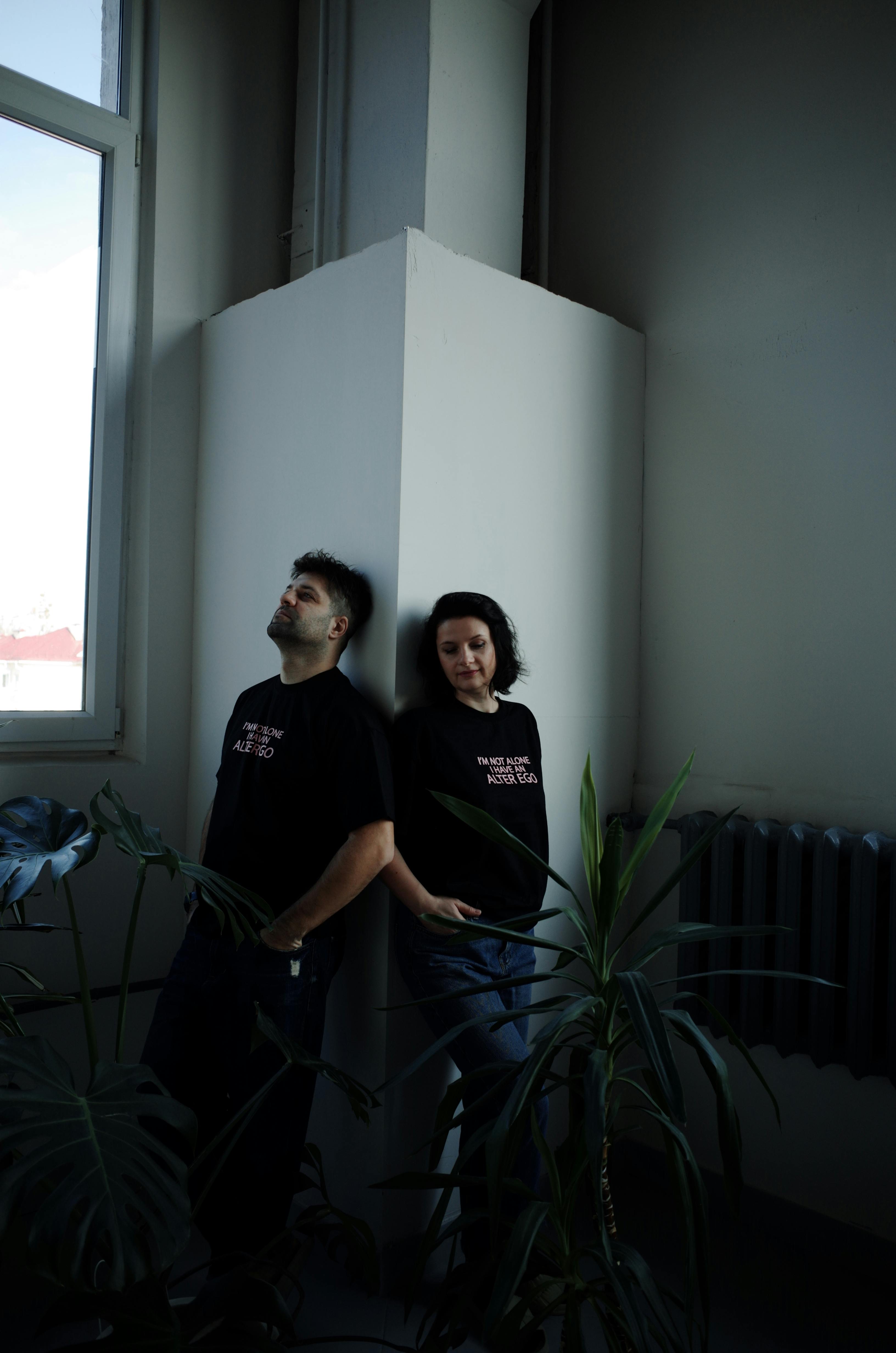 A man and woman leaning against a wall near houseplants, capturing a relaxed modern lifestyle indoors.