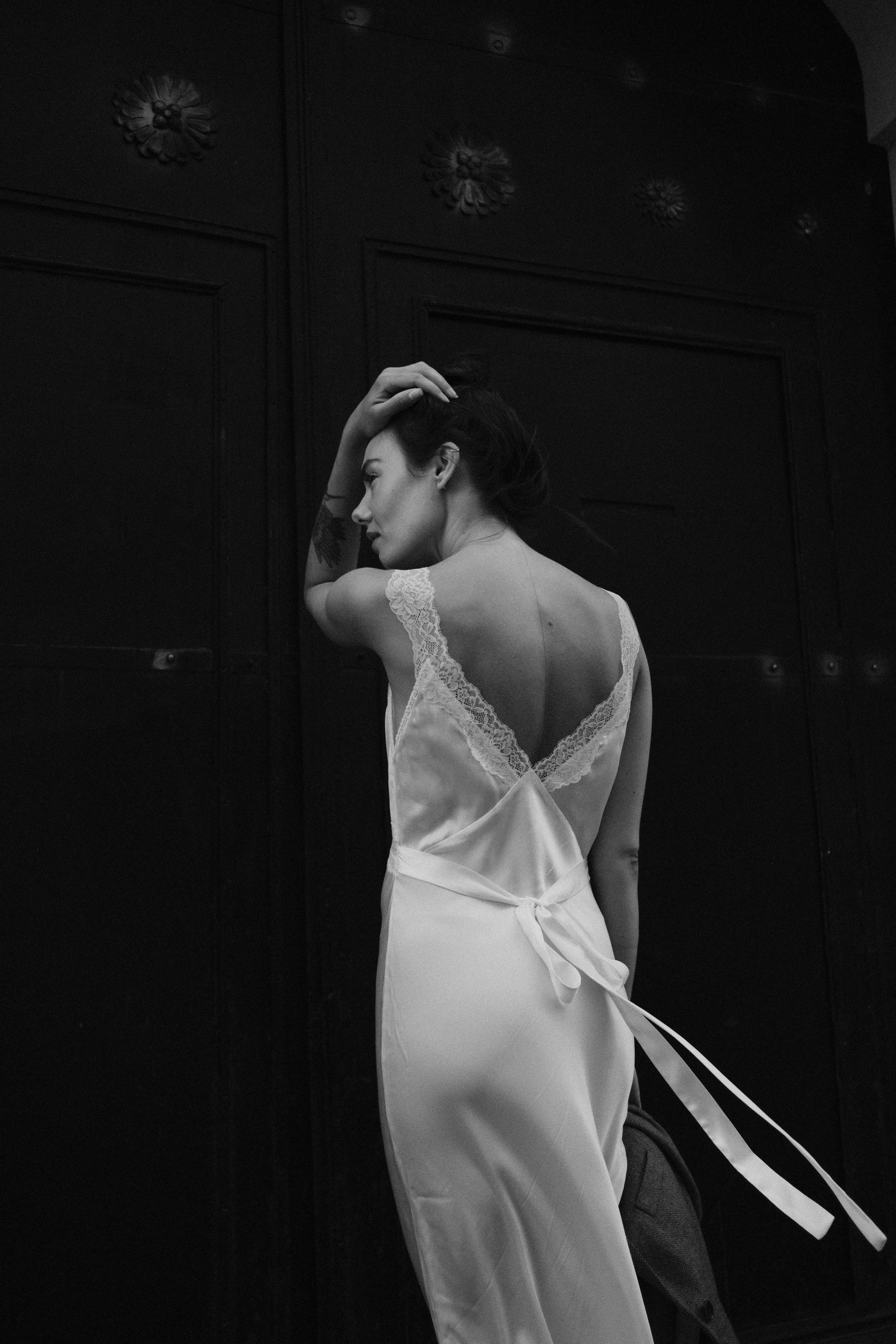 Black and white photo of a stylish woman in a delicate lace dress standing by a door.