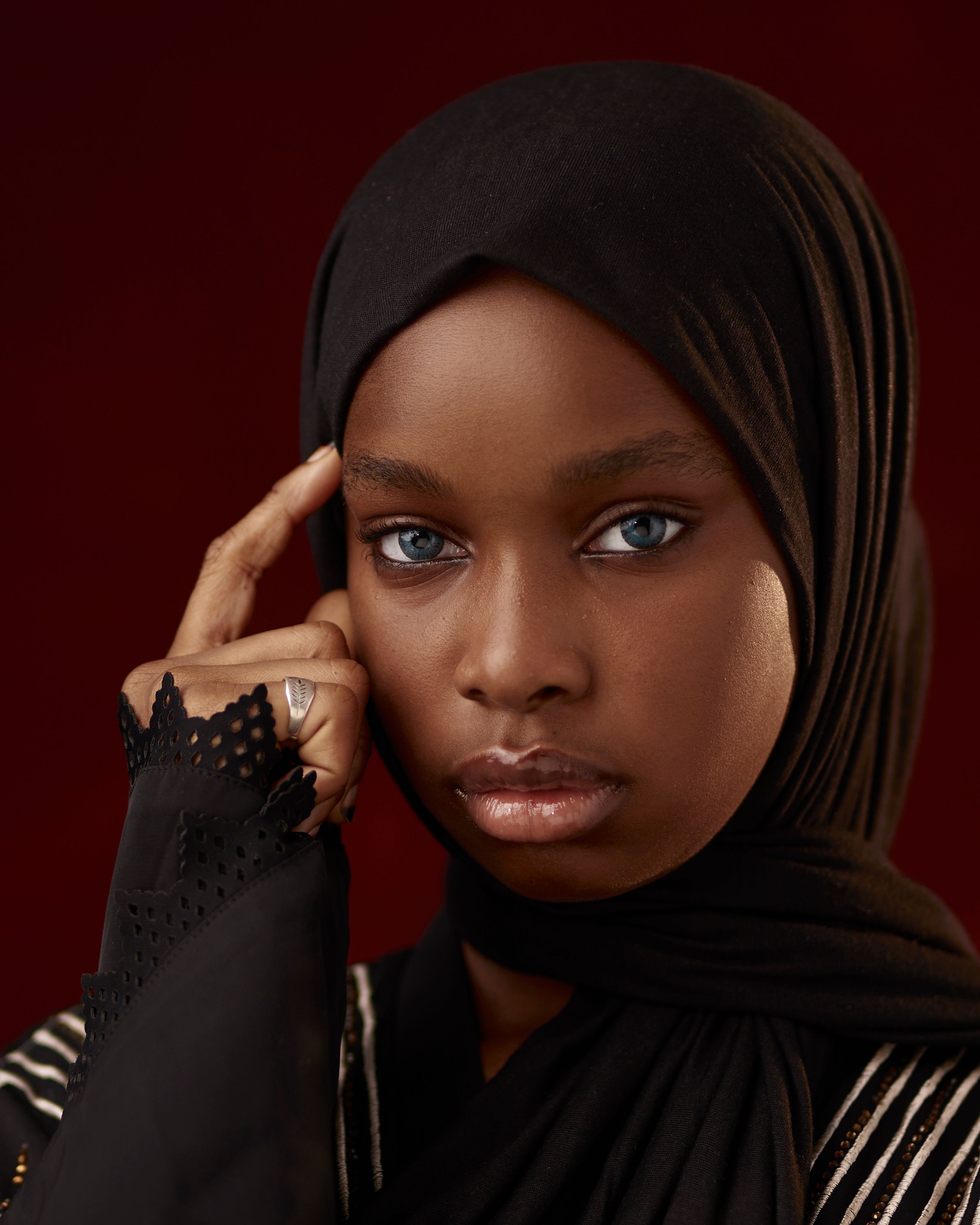 Striking studio portrait of a woman in a black hijab against a red backdrop.