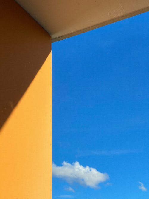 A blue sky and clouds are seen through a yellow wall