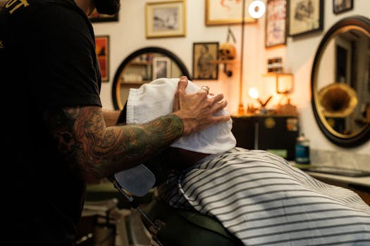 A barber with tattoo sleeves applies a hot towel in a vintage-style barbershop setting.