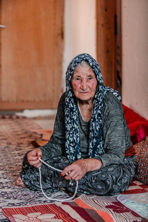 Free An elderly woman with a headscarf sits indoors, holding prayer beads on a patterned carpet. Stock Photo