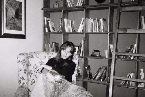 Free Woman sitting in an armchair in a library room, reading a book, surrounded by shelves and vintage decor. Stock Photo