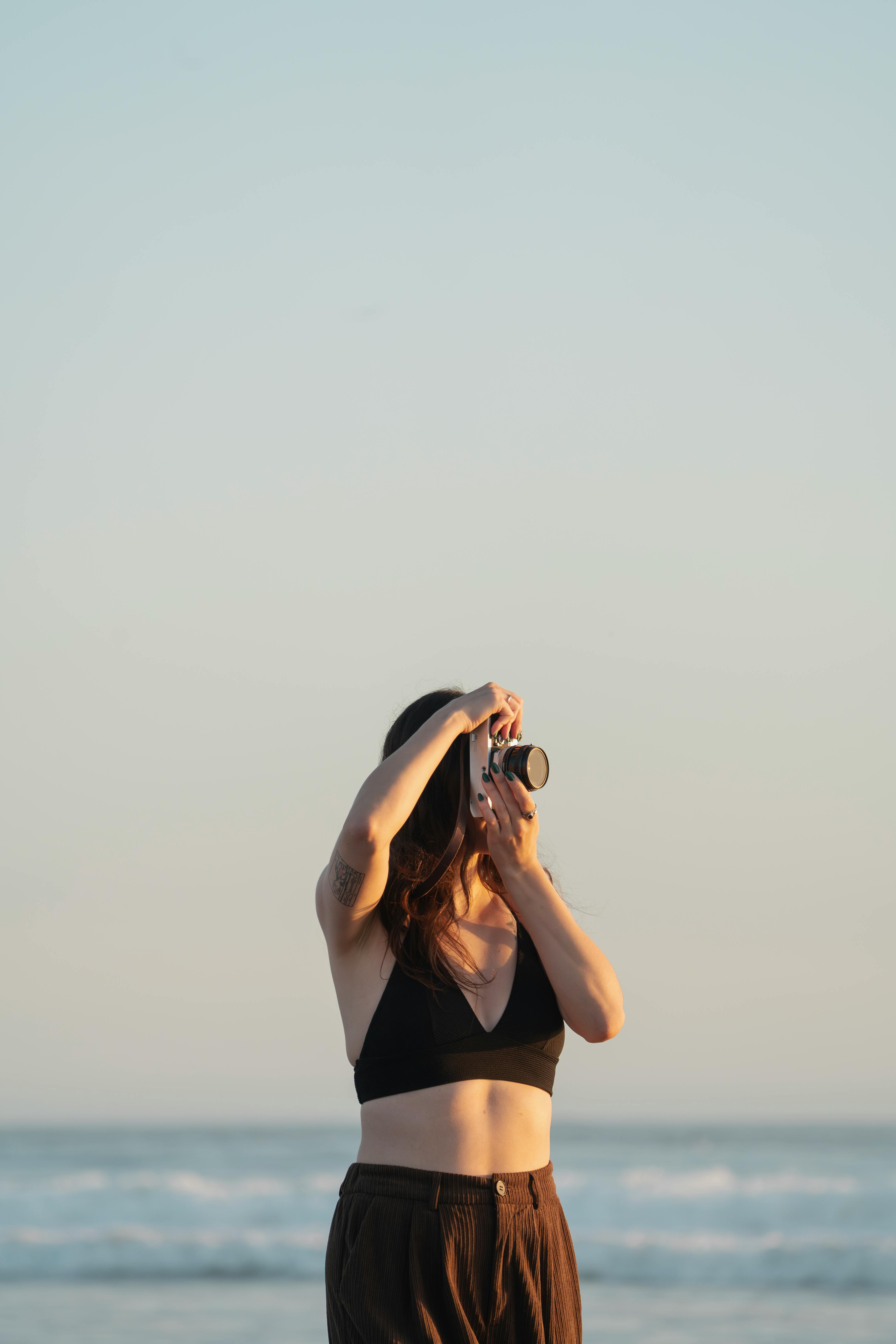 A woman in a black top stands on the beach taking photos at sunset.