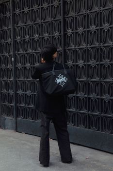 Man in black outfit standing against a modern urban wall holding a stylish bag.