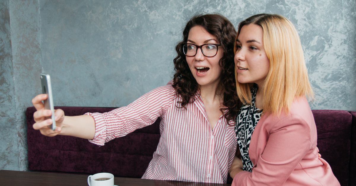 Two friends laughing together in a cozy coffee shop, one holding a phone but both engaged