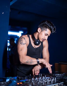 Young male DJ with tattoos mixing music in nightclub with headphones and sound mixer.