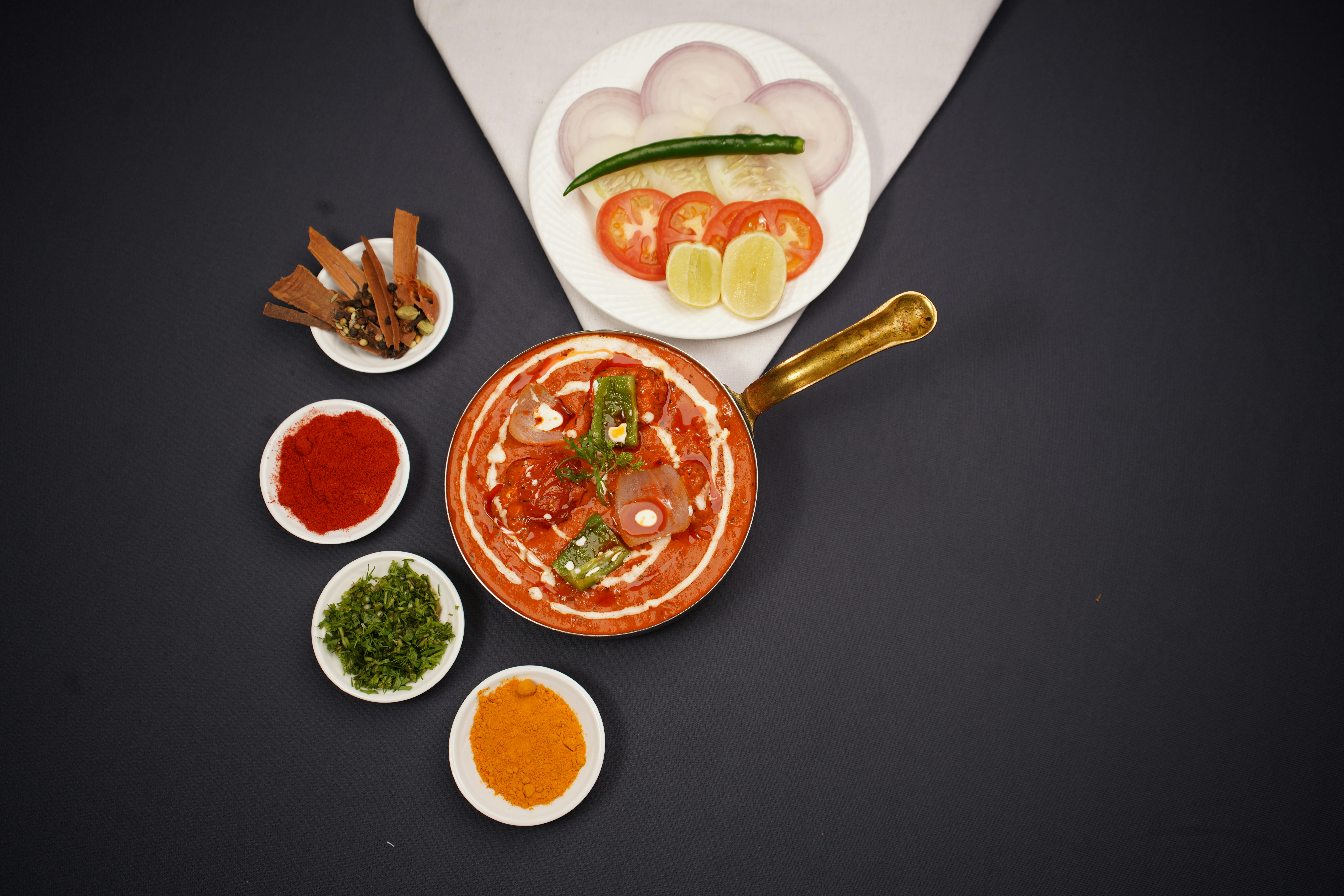 Top view of Indian curry in a pan with spices and fresh ingredients on a black background.