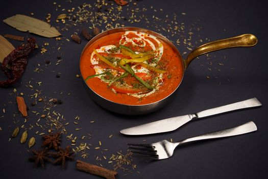 Close-up of a spicy red curry in a brass pot, garnished with herbs and surrounded by spices on a black background.