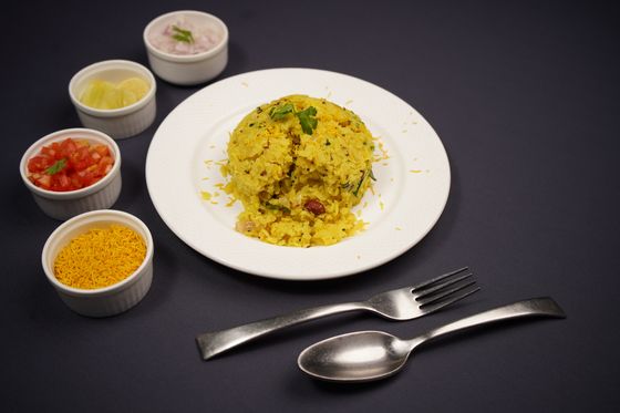 Mound of yellow poha on a plate with bowls of sev, tomato, onion and lemon