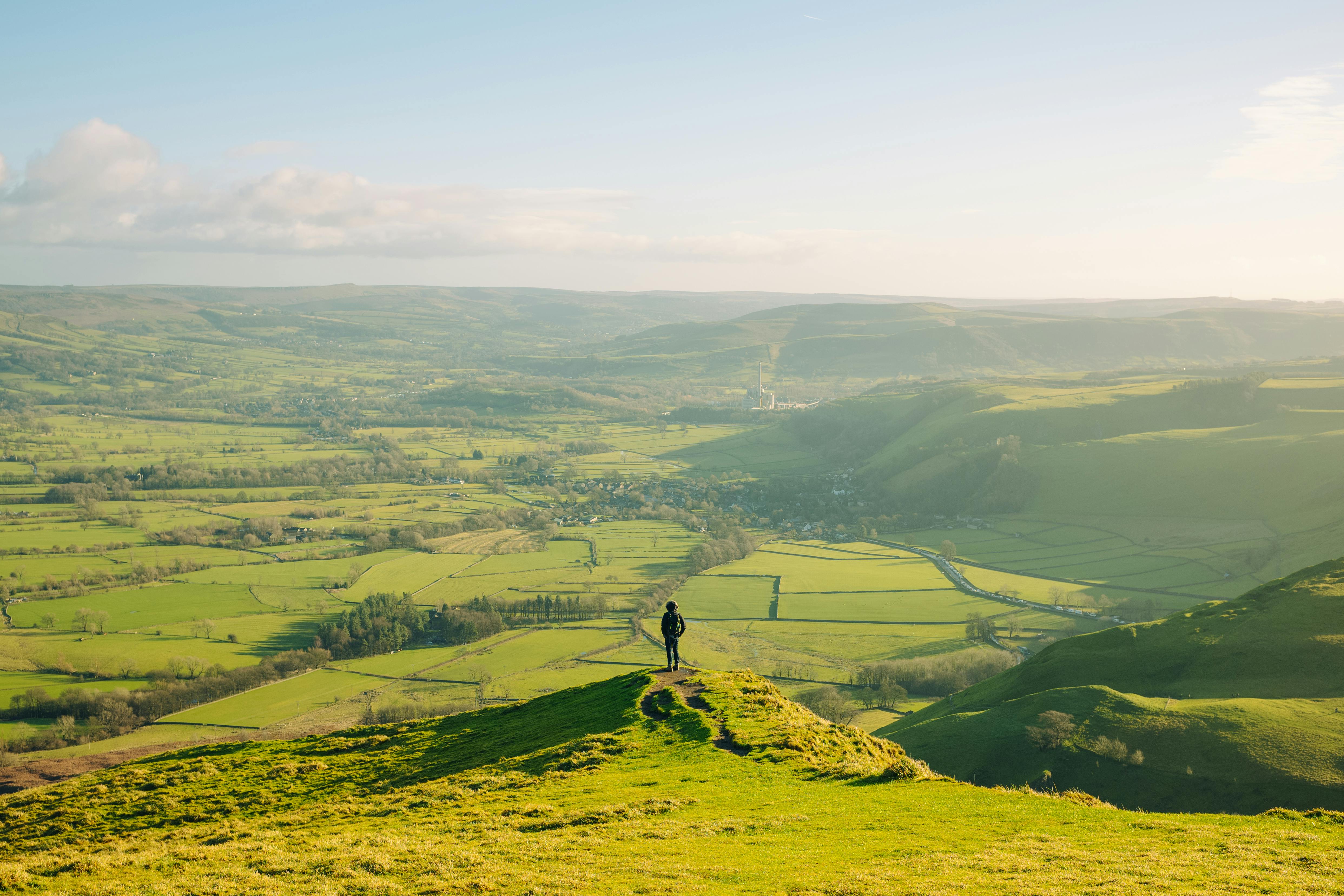 A lone hiker stands atop a hill, gazing over the vast green fields of England's Peak District under a clear sky.