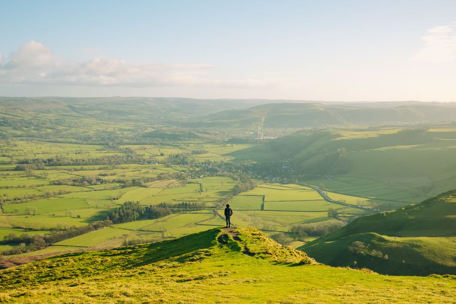 A lone hiker stands atop a hill, gazing over the vast green fields of England's Peak District under a clear sky.