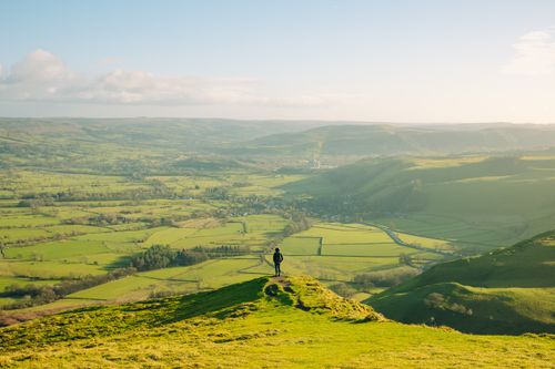Free A lone hiker stands atop a hill, gazing over the vast green fields of England's Peak District under a clear sky. Stock Photo