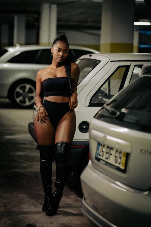 A woman in black thigh high boots and a black bodysuit standing next to a car