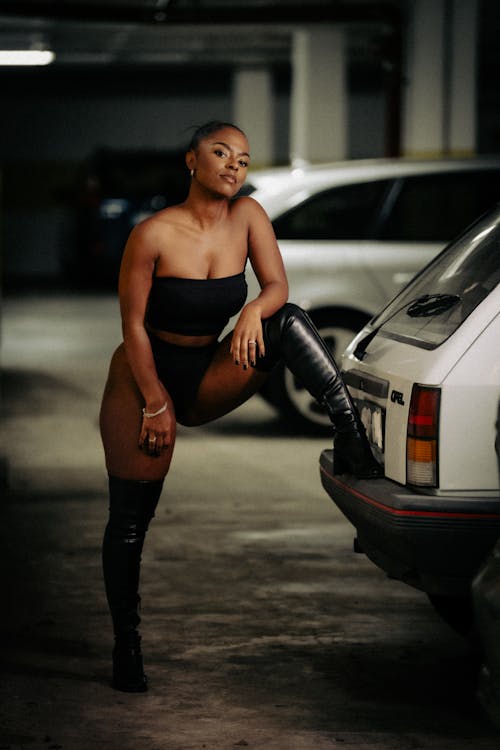 A woman in black thigh high boots and a black top leaning against a car