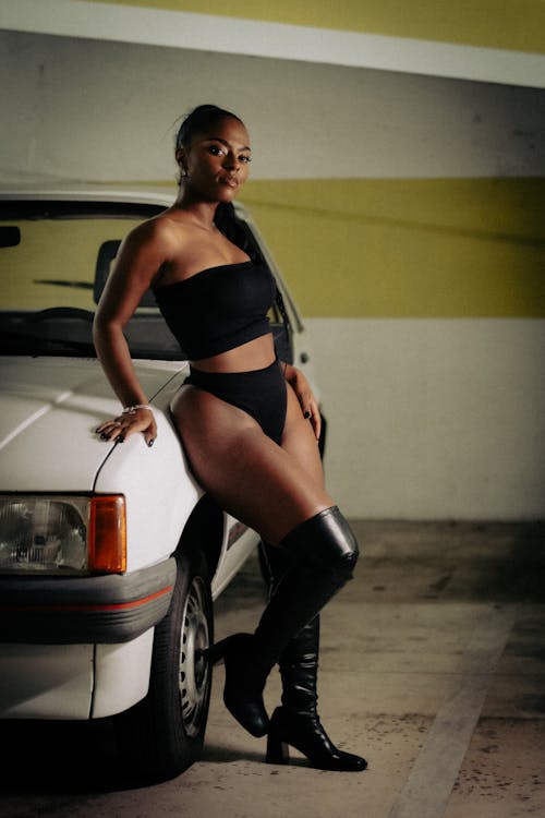 A woman in black thigh high boots and a black top posing next to a car