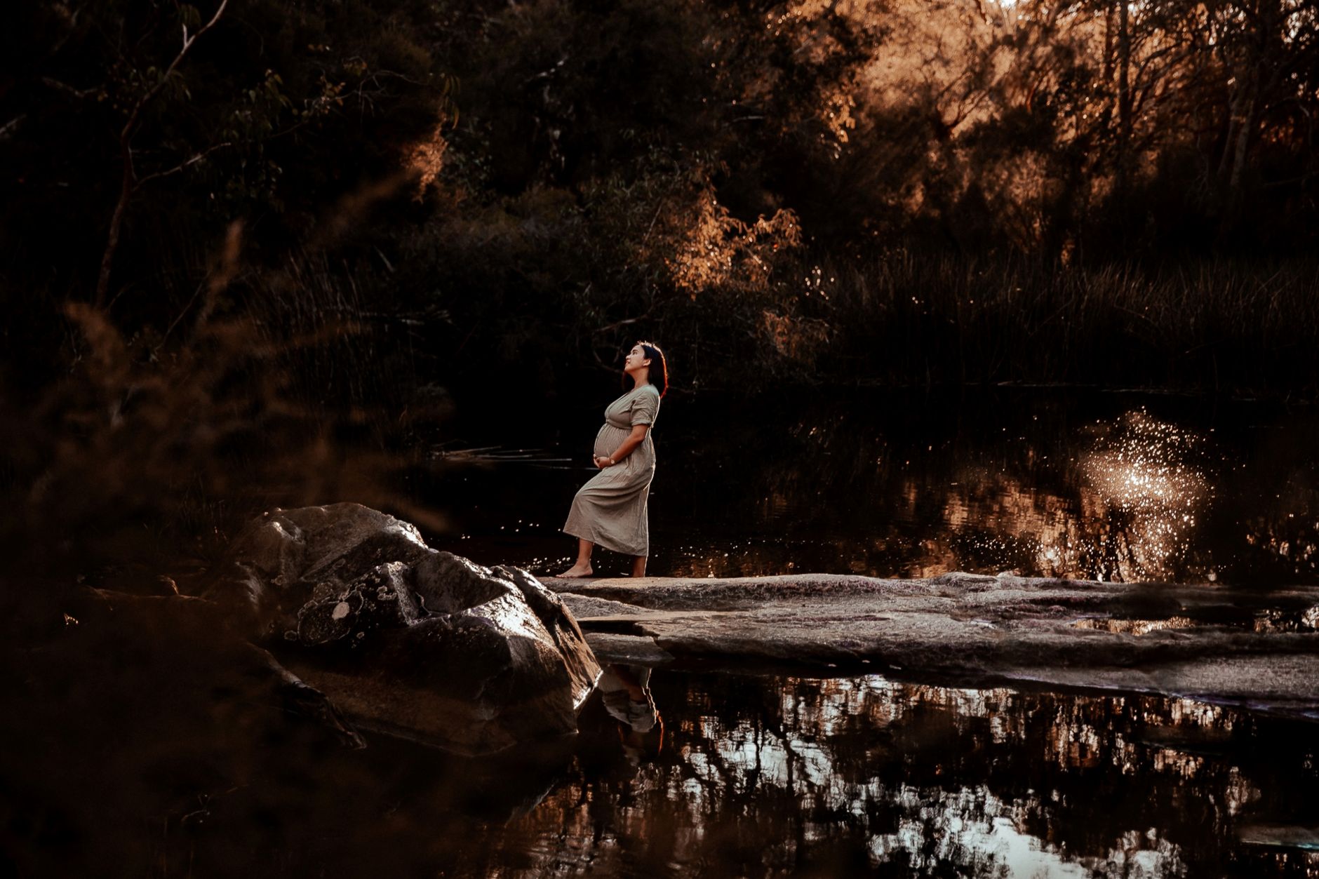 Pregnant woman in a serene forest setting by a reflective lake during sunset.