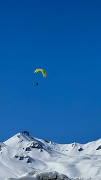 A paraglider flying over snowcapped peaks of the Caucasus Mountains against a clear blue sky.