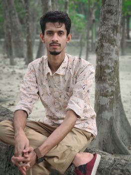 Casual portrait of a young man sitting on a tree trunk in a serene forest environment.