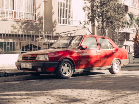 A vintage red sedan parked on a quiet city street in bright daylight.