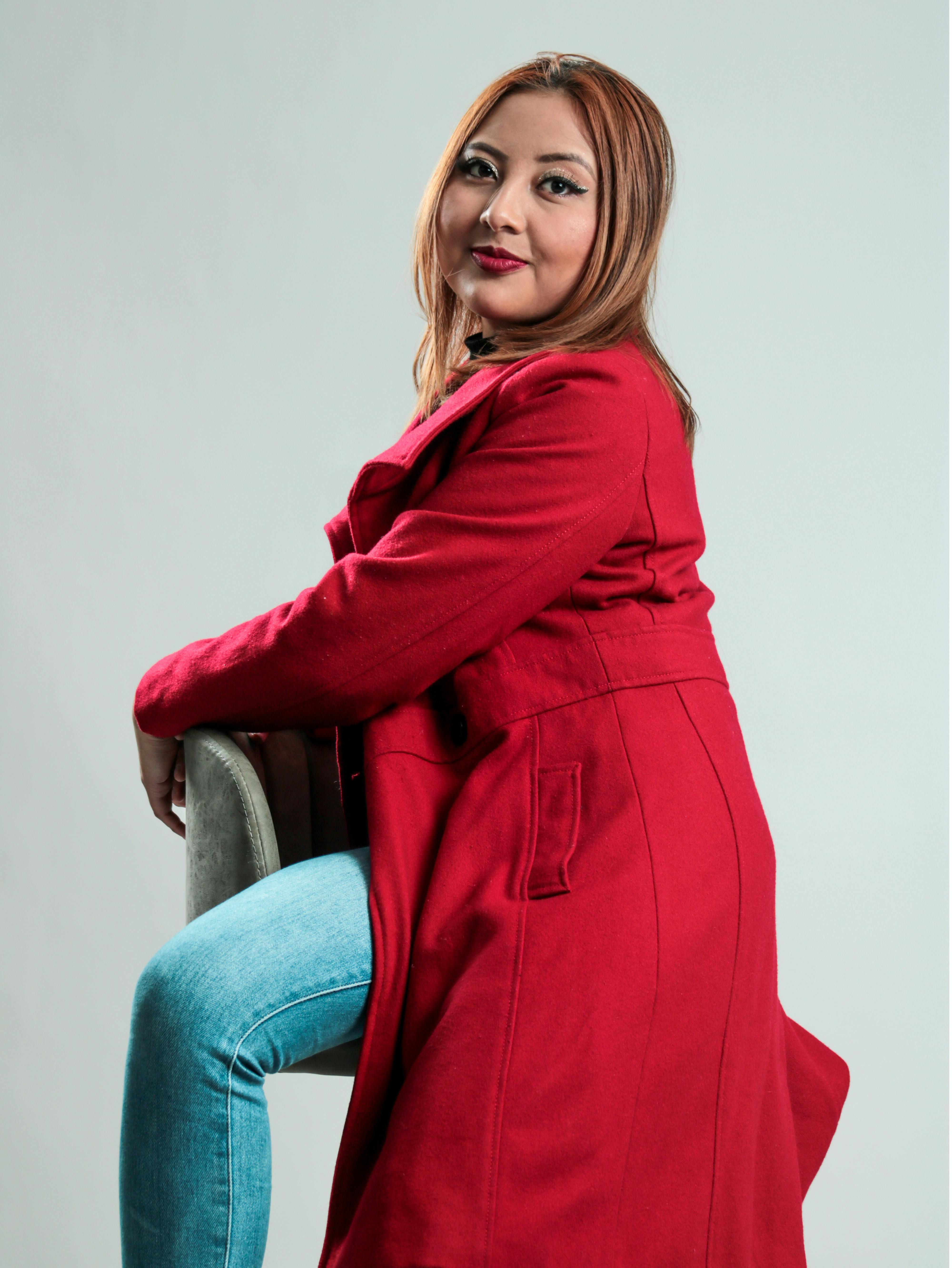 Portrait of a smiling woman in a red coat sitting on a chair in a studio setting.