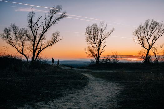 Two people walking along a rural path at sunset with bare trees silhouetted.