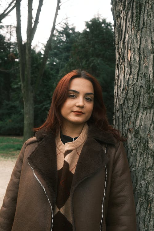 A woman in a brown coat and brown boots standing next to a tree