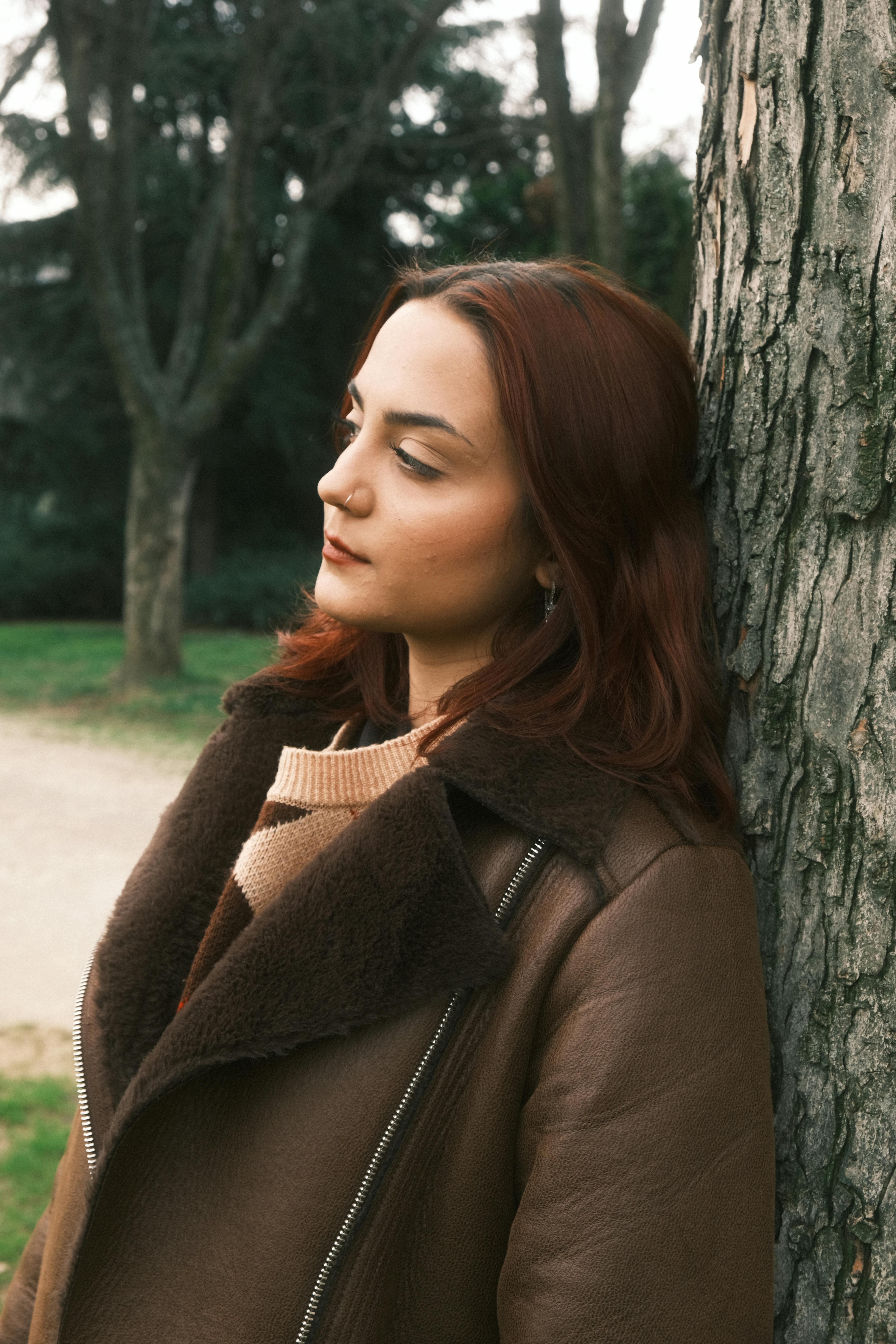 A fashionable woman with red hair leans against a tree, captured in an outdoor setting.