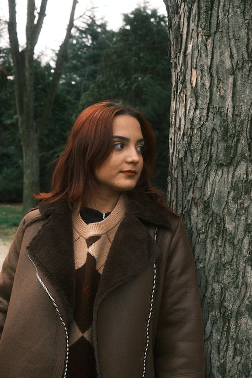 A woman with red hair and brown coat leaning against a tree