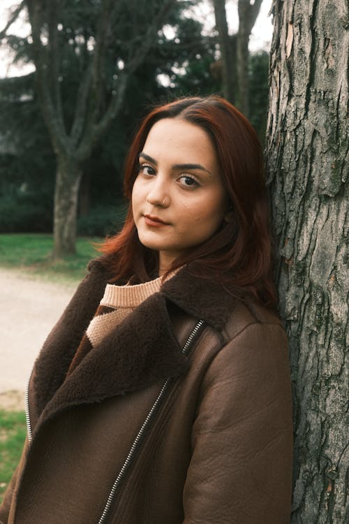 A woman in a brown coat leaning against a tree