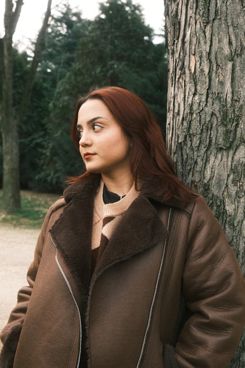 A woman in a brown coat leaning against a tree