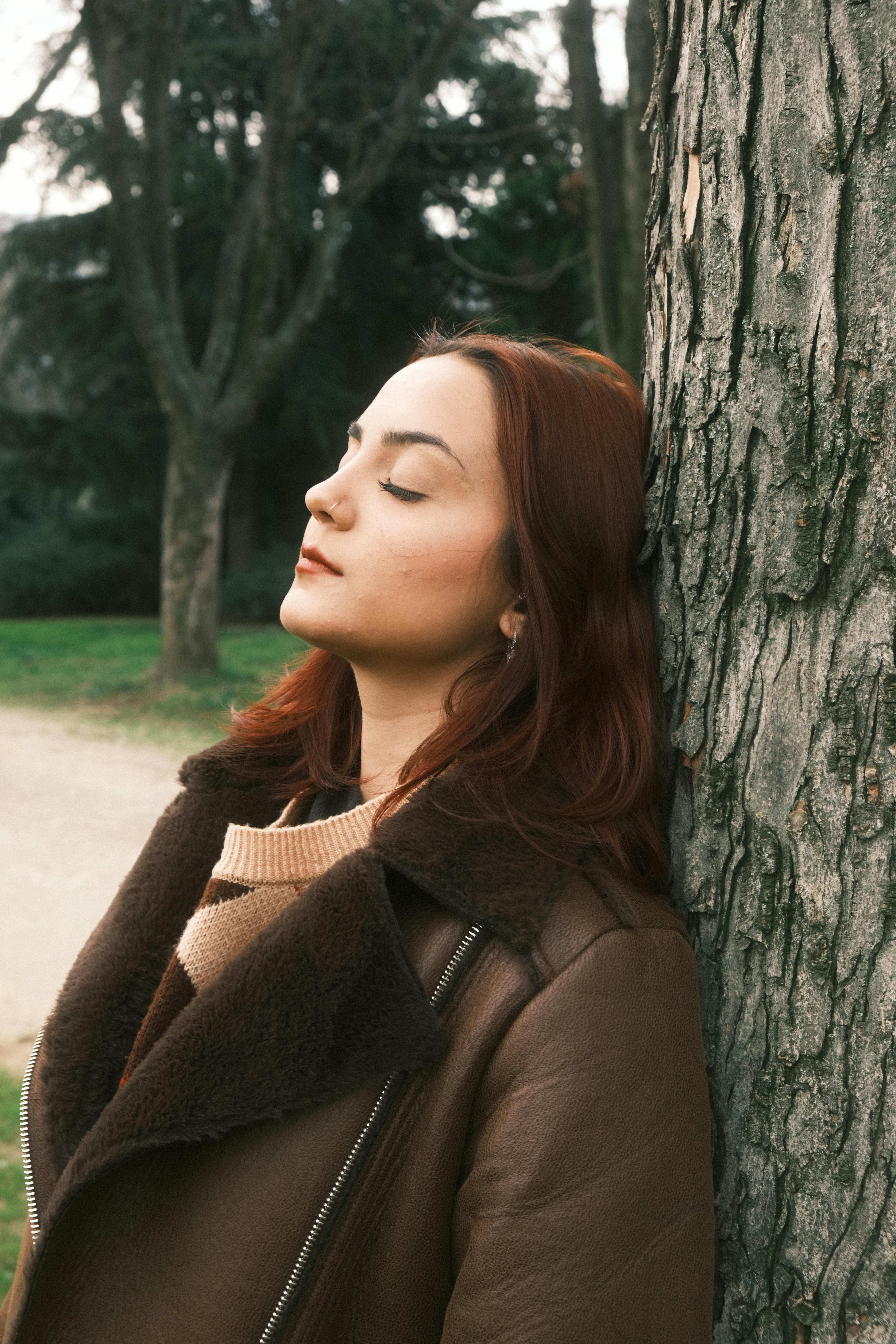Serene young woman with closed eyes meditates in a tranquil park setting, leaning against a tree for relaxation.