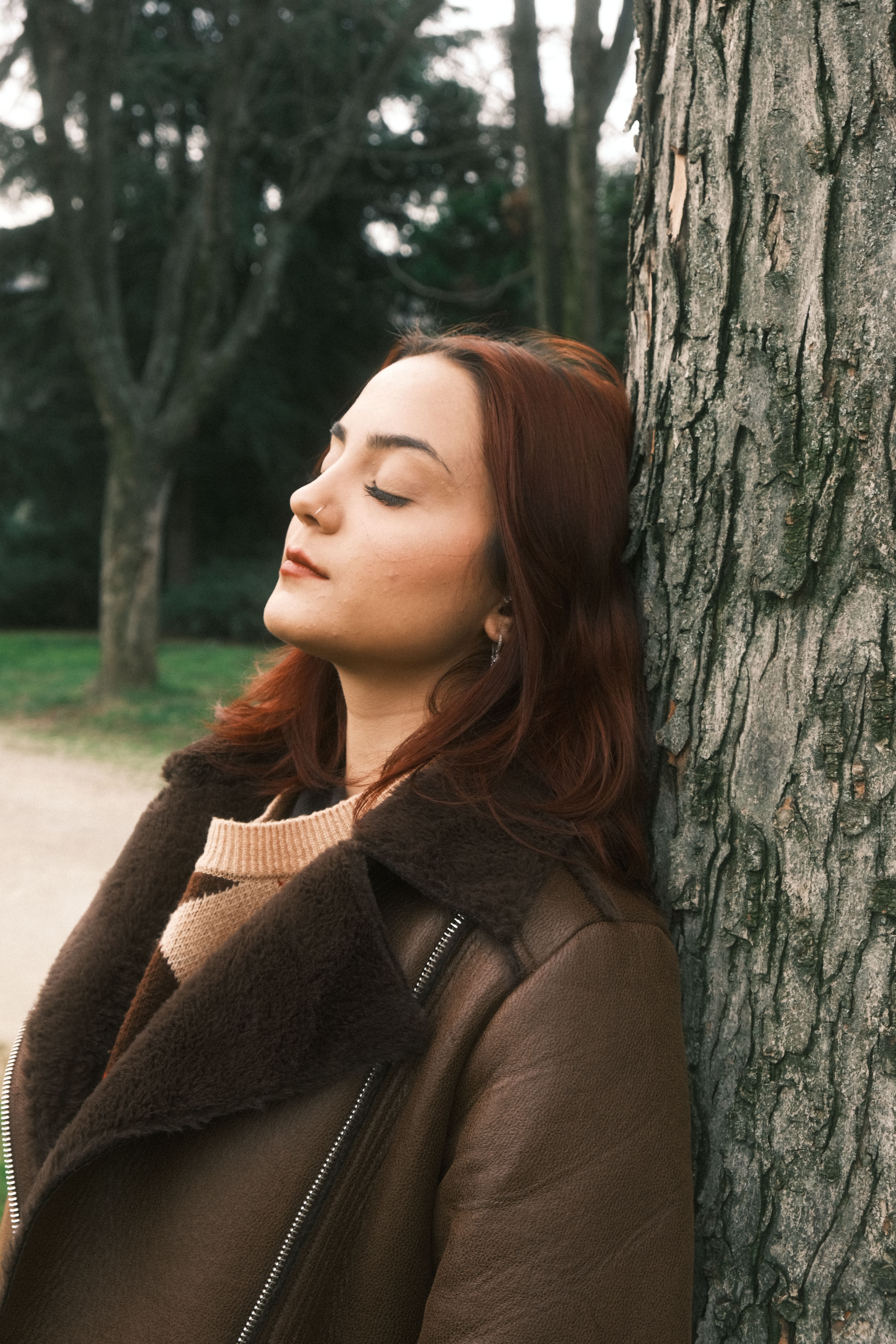 Woman with closed eyes in brown jacket leaning against a tree outdoors, capturing tranquility.