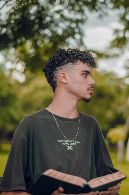 Young Man in a T-shirt Standing in a Park and Holding a Book 