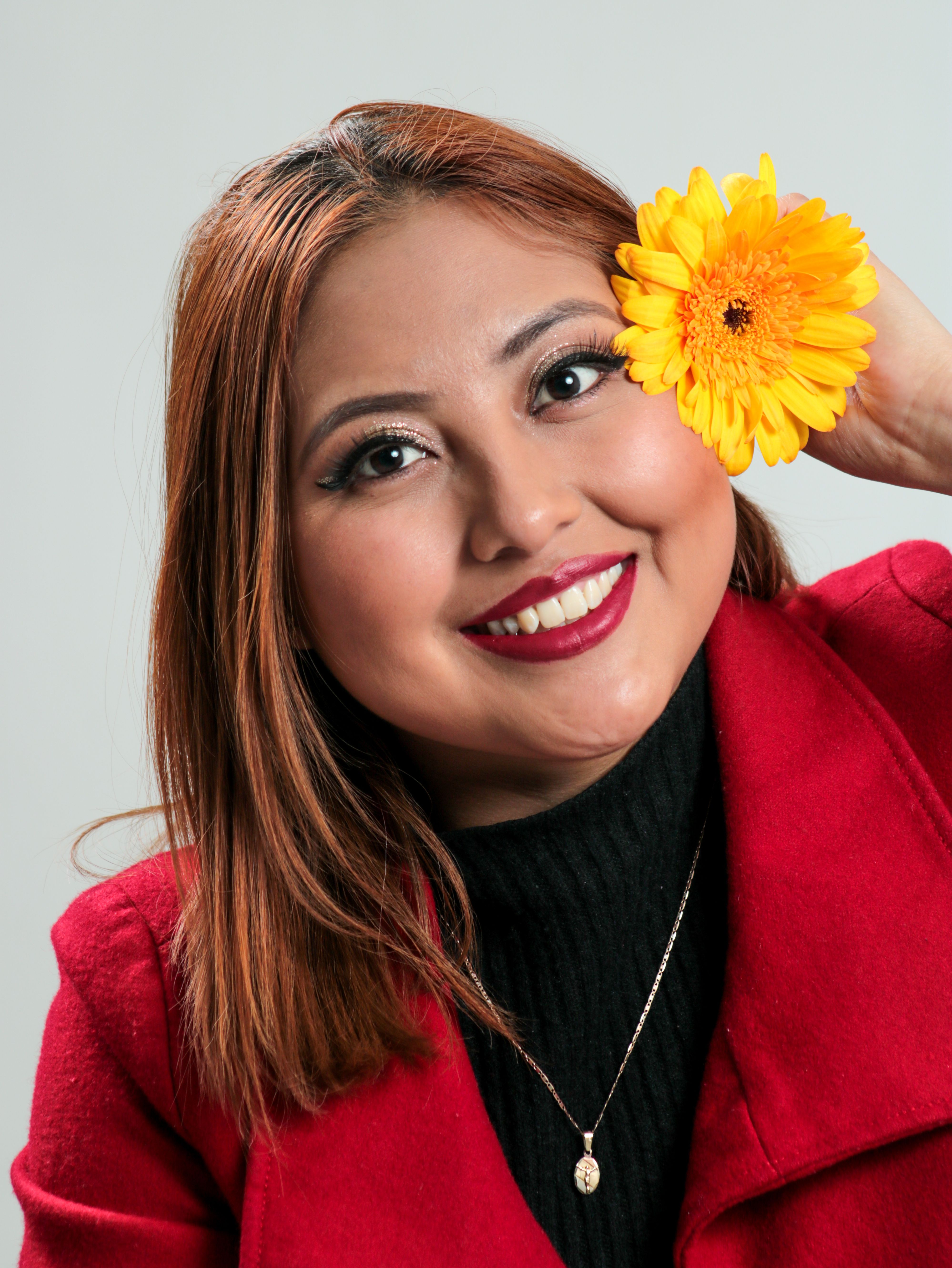 A cheerful woman in a red coat holds a yellow flower, showcasing elegance and beauty.