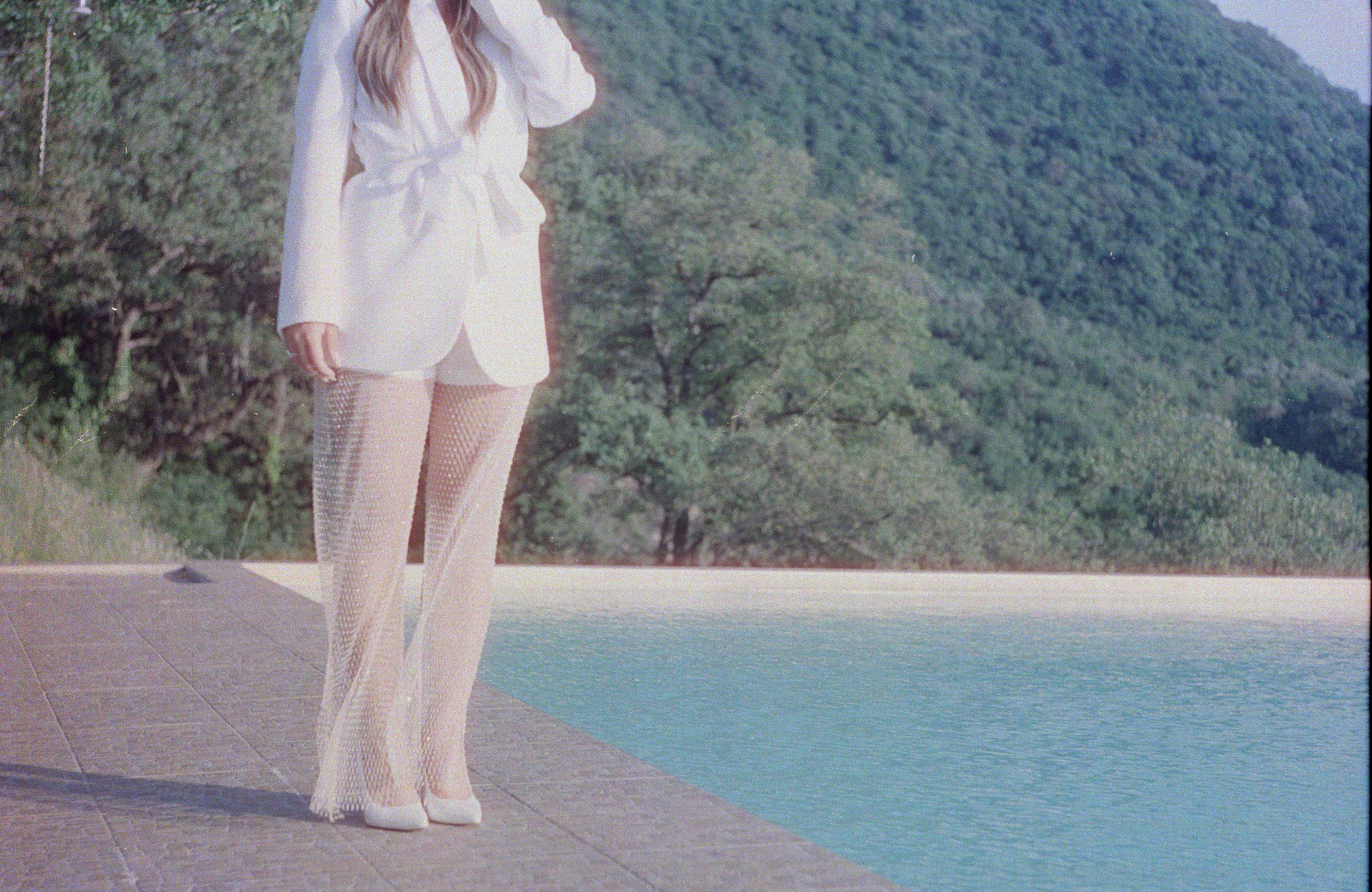 Chic fashion with a white blazer and mesh pants near a serene poolside setting.