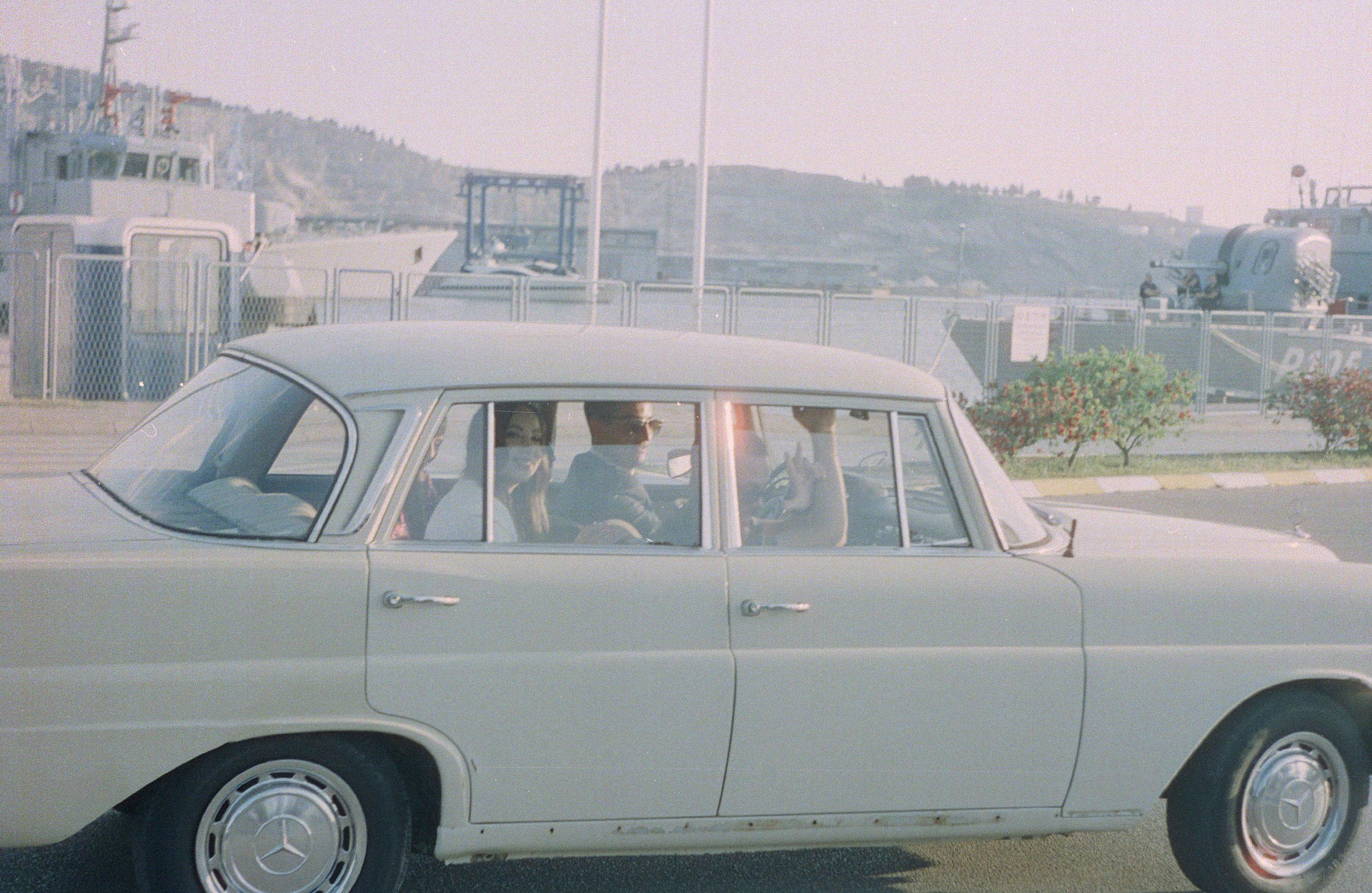 Classic white car with passengers near a coastal industrial setting.