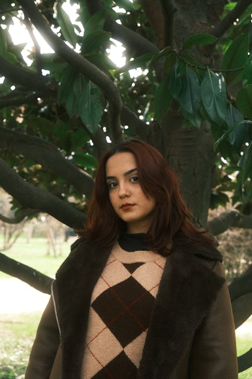 A woman in a sweater and jacket posing in front of a tree
