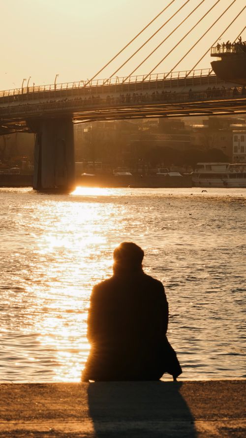Free A silhouette of a person sitting by the water, overlooking a bridge during sunset, evoking calmness and reflection. Stock Photo